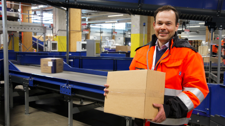 Michael Sundman seisoo pakettilajittelijan vieressä pitäen käsissään pakettia.